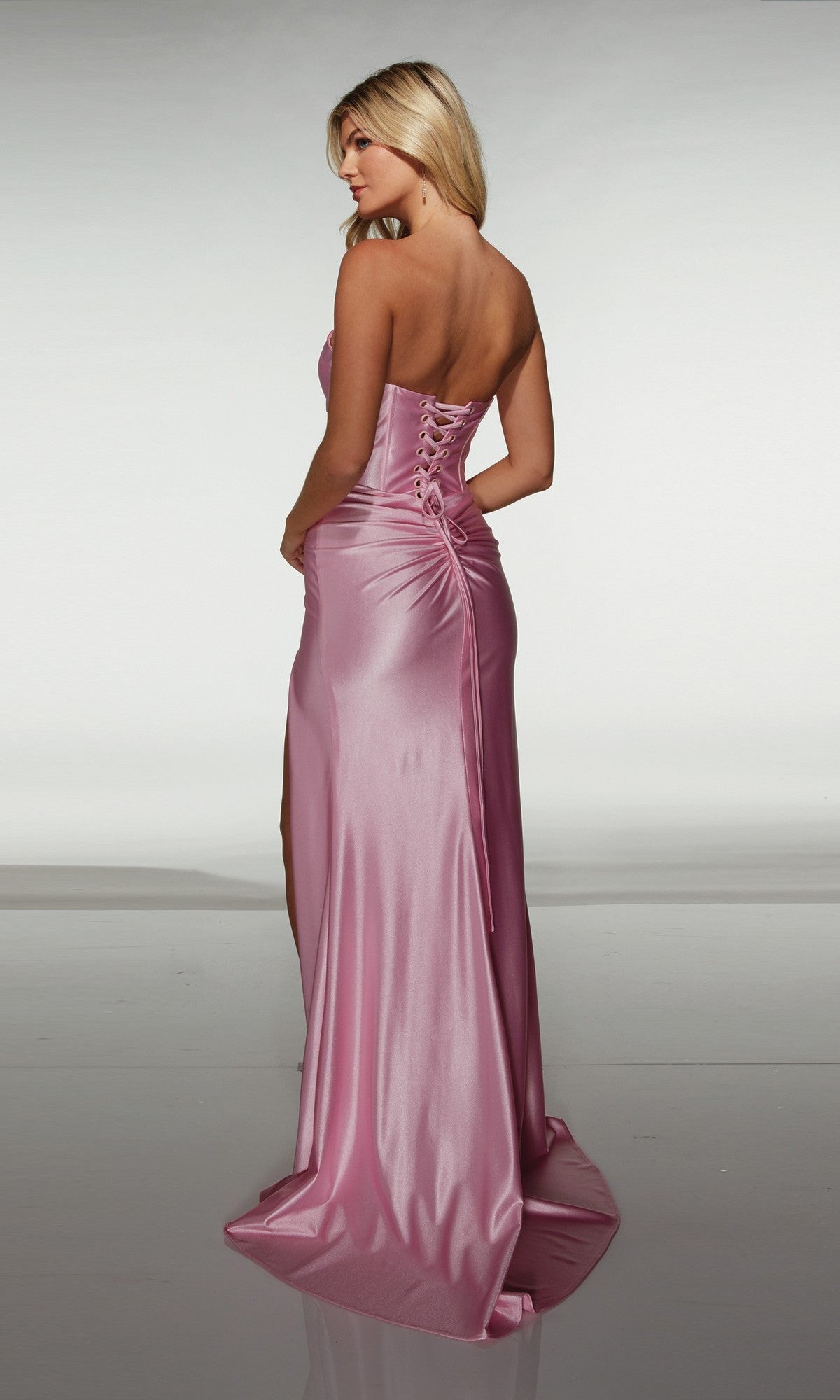 A woman wearing the Alyce 62046 long prom dress—strapless, floor-length pink satin with a fitted corset bodice and lace-up back—stands facing away from the camera.